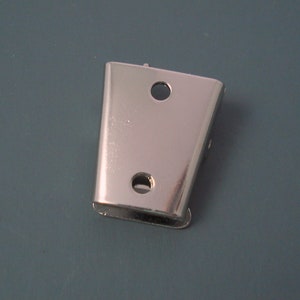 May include: A silver-colored metal hardware component with a trapezoidal shape, featuring two circular holes aligned vertically on its flat front surface. The piece has a smooth, reflective finish and a slightly curved profile along its side edges.