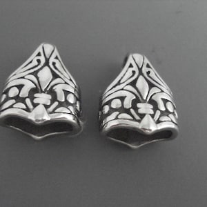 May include: Two silver-toned metal pendant charms with an intricate, ornate design. The charms are shaped like a pointed, upside-down teardrop with a decorative pattern.