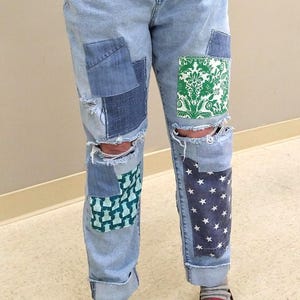 May include: Light blue denim jeans with various patches of different denim fabrics and patterns. The jeans have distressed areas at the knees and feature patches with green and white floral and star patterns.