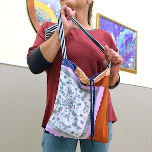 May include: A patchwork tote bag with a floral handkerchief design. The bag features a variety of fabrics in shades of white, pink, orange, and blue. The straps are striped in navy and white. The bag is held by a person.