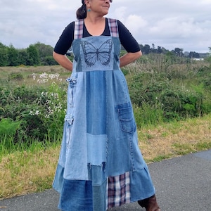 May include: A long denim patchwork dress featuring a black butterfly graphic on the chest, wide plaid shoulder straps, and multiple integrated pockets. The skirt is constructed from various shades of blue denim fabric panels with a plaid accent at the hem.