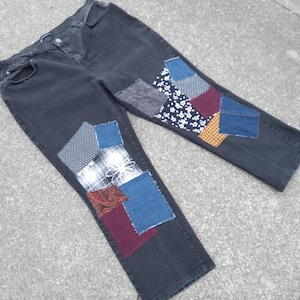 May include: Dark wash denim jeans with colorful patchwork details. Patches of various fabrics in shades of blue, red, white, and gold are sewn onto the knees. The jeans are laid flat on a gray surface.
