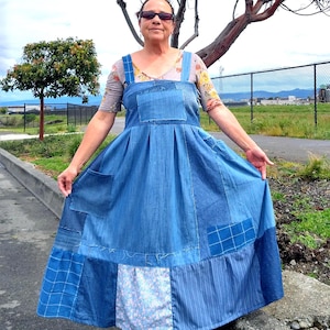 May include: A blue denim overall dress with patchwork details. The dress features a pleated skirt, large front pockets, and wide straps. The dress is worn over a floral print top.
