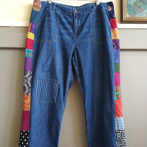 May include: Blue denim jeans with colorful patchwork accents down the sides. The patchwork includes various patterns and colors, such as polka dots, floral prints, and stripes. The jeans have a classic five-pocket design and are hanging on a hanger.