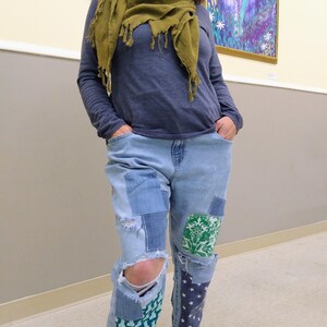 May include: A person wearing a blue long-sleeved top, a green scarf, and light blue jeans with patches and distressed areas. The jeans have patches with green and white floral patterns and blue star patterns. Brown shoes complete the outfit.