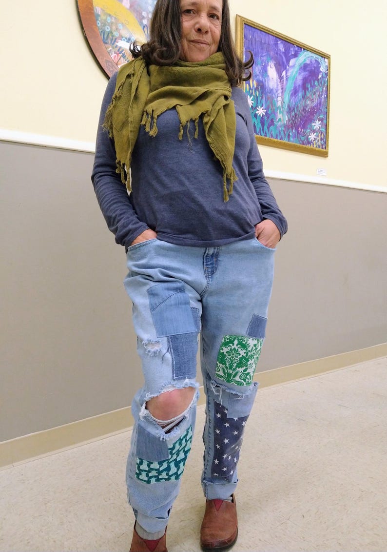 May include: A person wearing a blue long-sleeved top, a green scarf, and light blue jeans with patches and distressed areas. The jeans have patches with green and white floral patterns and blue star patterns. Brown shoes complete the outfit.