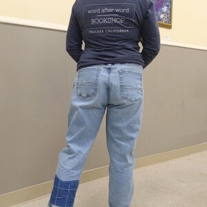 May include: A person wearing a navy blue long-sleeved top with the text "word after word BOOKSHOP TRUCKEE CALIFORNIA" and light blue jeans. The jeans have a blue patch on the left leg and are cuffed. Brown shoes are visible.