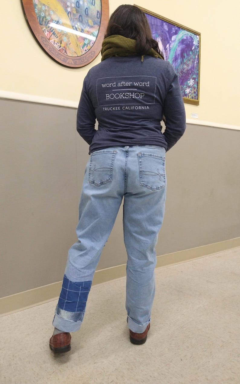 May include: A person wearing a navy blue long-sleeved top with the text "word after word BOOKSHOP TRUCKEE CALIFORNIA" and light blue jeans. The jeans have a blue patch on the left leg and are cuffed. Brown shoes are visible.