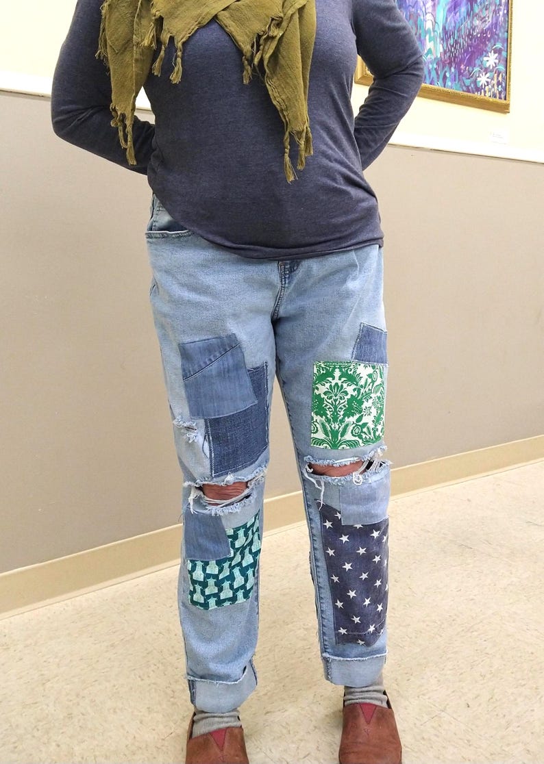 May include: Light blue denim jeans with various patches of different denim fabrics and patterns. The jeans have distressed areas at the knees and feature patches with green and white floral and star patterns.