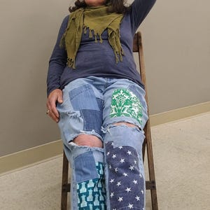 May include: Light blue denim jeans with patchwork designs, including a green floral pattern, a blue geometric pattern, and a navy blue star pattern. The jeans have distressed details and ripped knees. The person is wearing a blue long-sleeve shirt and a green scarf.