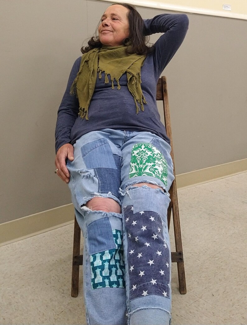 May include: Light blue denim jeans with patchwork designs, including a green floral pattern, a blue geometric pattern, and a navy blue star pattern. The jeans have distressed details and ripped knees. The person is wearing a blue long-sleeve shirt and a green scarf.