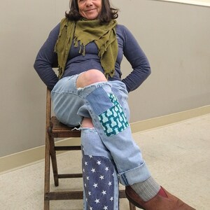 May include: Light blue denim jeans with patches of blue fabric and white stars. The jeans have ripped knees and are cuffed at the ankles. The person is wearing a blue long-sleeve shirt, a green scarf, and brown shoes.