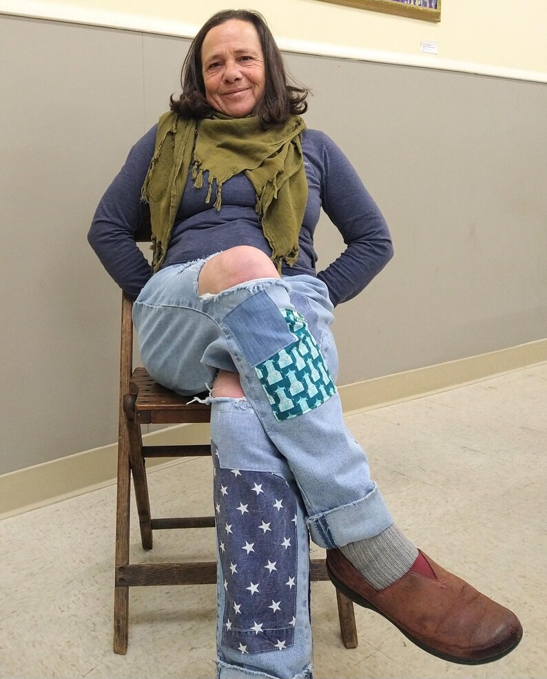 May include: Light blue denim jeans with patches of blue fabric and white stars. The jeans have ripped knees and are cuffed at the ankles. The person is wearing a blue long-sleeve shirt, a green scarf, and brown shoes.