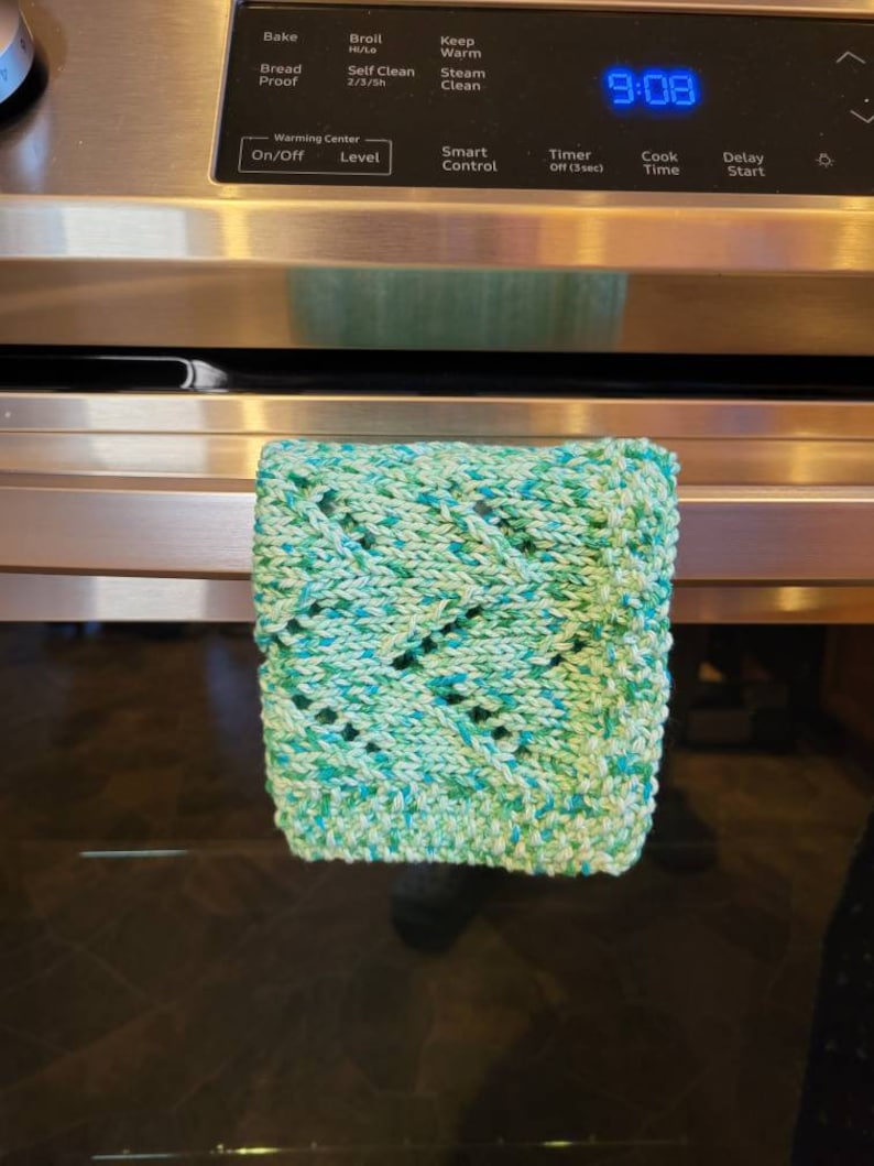 May include: A green and blue knitted dishcloth hangs on the handle of a stainless steel oven. The oven has a digital display showing the time 9:08.