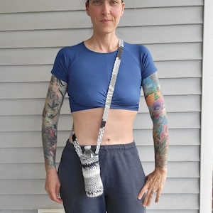 May include: A person wearing a blue cropped top and gray sweatpants with a crocheted water bottle holder slung over their shoulder. The holder is black, white, and gray with a clear plastic water bottle inside.