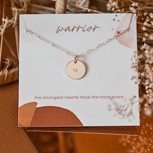 May include: A delicate gold-colored necklace with a circular pendant featuring a small heart. The necklace is displayed on a white card with the word "warrior" and the phrase "the strongest hearts have the most scars".