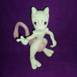 May include: A green and pink plush toy of the Pokemon character Mewtwo. The toy is standing on a purple surface.