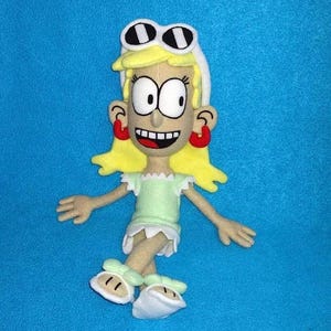 May include: A handmade plush doll of a cartoon character with blonde hair, large eyes, and a wide smile. The doll wears a light green dress, white shoes, and red earrings. The doll has white sunglasses on its head.