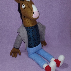Puede incluir: Un peluche de un personaje con cabeza de caballo, con una chaqueta gris, un su&eacute;ter azul con un patr&oacute;n azul oscuro y pantalones azul claro. El juguete tiene manos marrones y una cabeza de caballo marr&oacute;n con una mancha blanca y hocico rosa. Zapatos rojos y blancos completan el look.