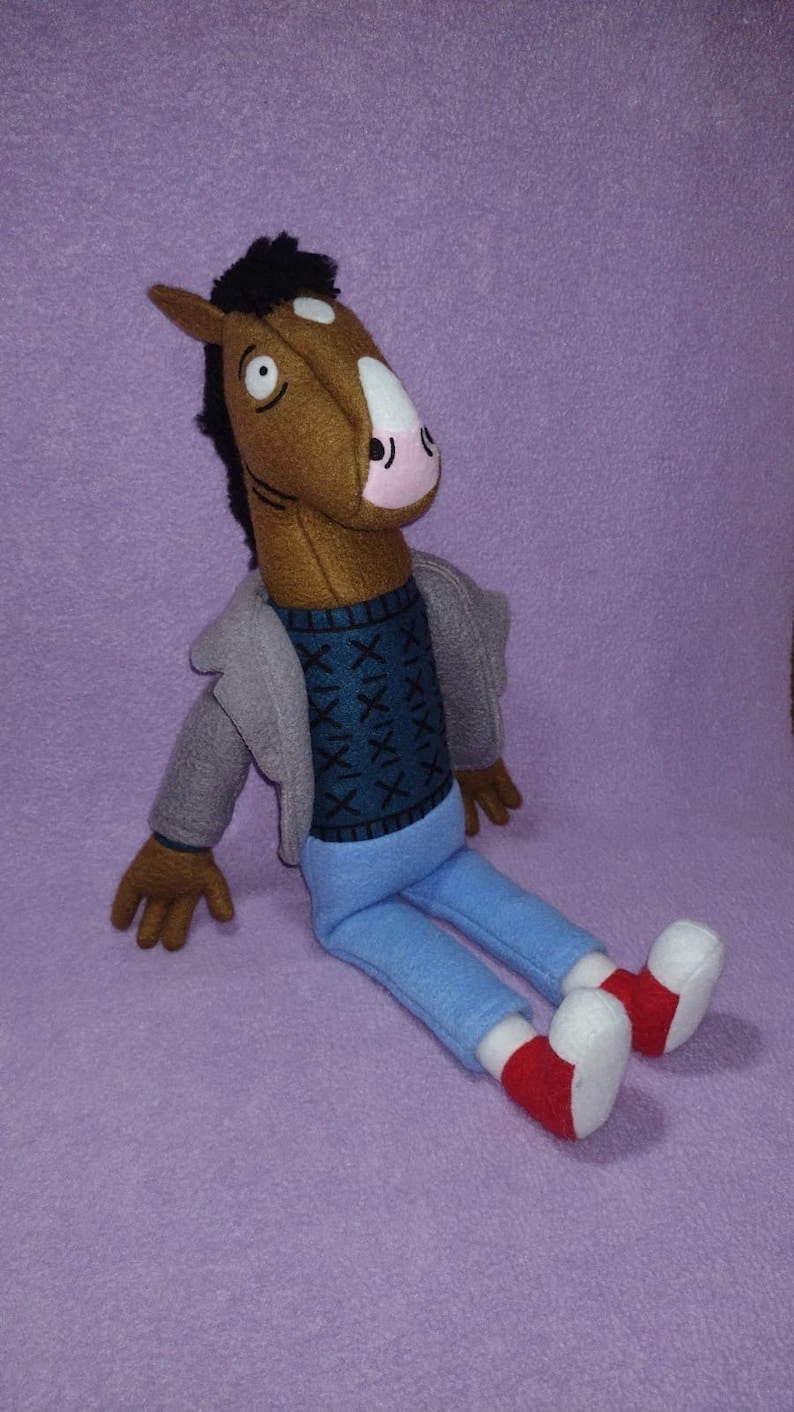 Puede incluir: Un peluche de un personaje con cabeza de caballo, con una chaqueta gris, un su&eacute;ter azul con un patr&oacute;n azul oscuro y pantalones azul claro. El juguete tiene manos marrones y una cabeza de caballo marr&oacute;n con una mancha blanca y hocico rosa. Zapatos rojos y blancos completan el look.