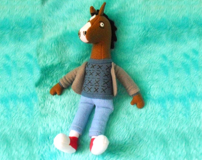 Puede incluir: Un juguete de peluche hecho a mano con forma de caballo con cabeza marr&oacute;n, melena negra y hocico blanco. El caballo lleva una chaqueta gris sobre un su&eacute;ter azul con un patr&oacute;n de X, pantalones azul claro y zapatos blancos con detalles rojos. El juguete est&aacute; sobre un fondo turquesa.