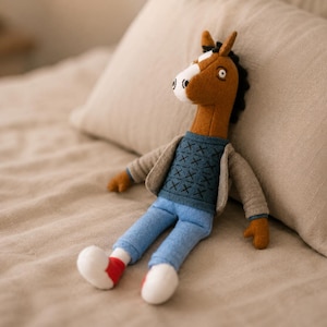 Puede incluir: Un muñeco de peluche con forma de caballo, cabeza marrón, hocico blanco y crin negra. Viste un suéter azul con un patrón de punto de cruz azul oscuro, una chaqueta abierta de color beige claro, pantalones azul claro y zapatos blancos con detalles rojos.