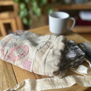 以下が含まれることがあります： ピンク、グレー、ベージュの色合いの複雑な刺繍が施されたオフホワイトの生地で作られた巾着袋。バッグはギャザーの寄せられた開口部に長い共布の紐が付いており、白黒の表紙の長方形の本が入っています。