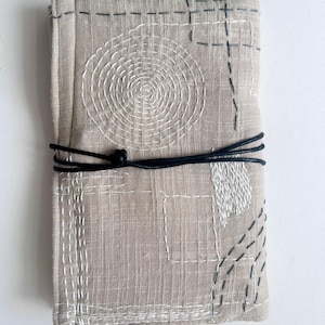 Peut inclure: Une pochette rectangulaire en tissu beige ornée de motifs géométriques cousus en blanc et gris foncé, incluant une spirale et des formes rectangulaires. Un cordon noir fin est noué au centre. Le texte indique : MY VERSION OF THE RETURNING KEEPER WRAP. YOURS WILL BE UNIQUE TO YOU AND YOUR COMPANION.