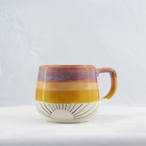 May include: A ceramic mug with a sunrise design. The mug features horizontal bands of color, including shades of purple, orange, and yellow. The bottom half of the mug is white with a sunburst pattern. The handle is a matching color.