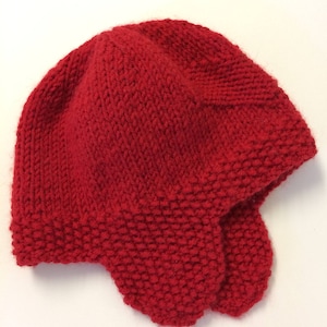 May include: A red knitted hat with ear flaps. The hat is made with a simple stitch pattern and has a rounded crown.