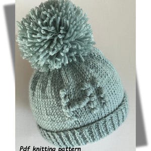 May include: A knitted beanie in a soft teal color with a large pom-pom on top. The hat features a ribbed brim and a textured pattern on the crown. The text "Pdf knitting pattern" is visible at the bottom.