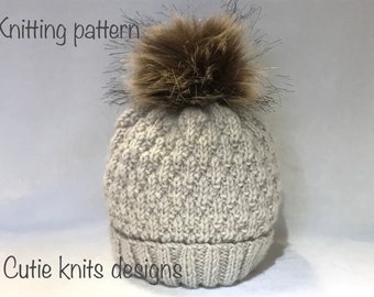 Könnte beinhalten: Strickmuster für eine beige Strickmütze mit einem braunen Kunstfellbommel. Die Mütze hat einen gerippten Rand und ein strukturiertes Strickmuster. Der Text "Cutie knits designs" ist am unteren Rand des Bildes sichtbar.