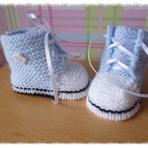 Könnte beinhalten: Ein Paar gestrickte Babyschuhe in Blau und Weiß mit weißen Satinband-Schnürsenkeln. Die Schuhe haben einen schwarzen Besatz um die Sohle.