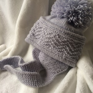 Op de afbeelding: Een grijze gebreide muts met een pompon bovenop en een bijpassende sjaal. De muts en sjaal zijn gemaakt van een zacht, getextureerd garen.