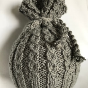 May include: A gray knitted drawstring bag with a textured cable knit pattern. The bag is made of soft yarn and has a gathered top with a knitted cord closure. The bag is suitable for small gifts or storage.