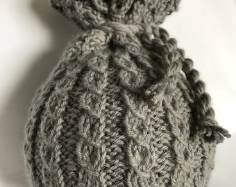 May include: A gray knitted drawstring bag with a textured cable knit pattern. The bag is made of soft yarn and has a gathered top with a knitted cord closure. The bag is suitable for small gifts or storage.