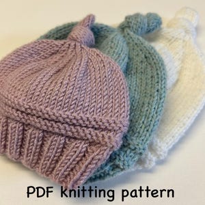 May include: Three knitted baby hats in pink, blue and white with a knotted top. PDF knitting pattern.