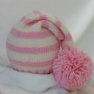 Op de afbeelding: Een gebreide baby muts met roze en witte strepen en een roze pompon.