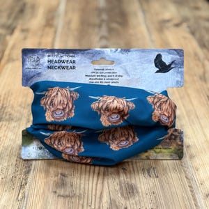 Könnte beinhalten: Blaues multifunktionales Headwear-Neckwear mit einem sich wiederholenden Muster von Highland-Kuhgesichtern. Die Verpackung enthält Text, der auf UPF30-Sonnenschutz, feuchtigkeitsableitende Eigenschaften und Atmungsaktivität hinweist. Eine schwarze Vogelgrafik ist ebenfalls sichtbar.