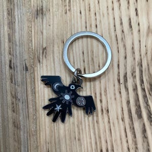 May include: A silver keyring with a black bird-shaped charm. The charm has a stylized bird design with white and grey accents, including a moon, star, and gear-like shapes. The keyring is on a wooden surface.