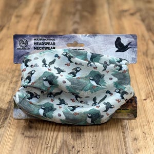 May include: Multifunctional headwear and neckwear featuring a pattern of puffins in flight and on the ground, set against a light green and grey landscape. The packaging reads "Multifunctional Headwear Neckwear".