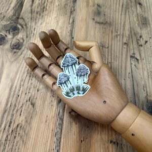 May include: A sticker depicting three stylized mushrooms with gray caps and white stems. The mushrooms have a dripping effect, and are set against green grass. The sticker is held by a wooden hand.