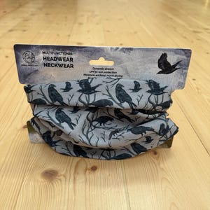 Puede incluir: Un tubo de cuello multifuncional de color gris claro con un patrón repetitivo de cuervos negros posados en ramas desnudas. El empaque incluye el texto Rona Innes Art, Multifunctional Headwear Neckwear, Dynamic stretch, UPF30 sun protection, Moisture-wicking.