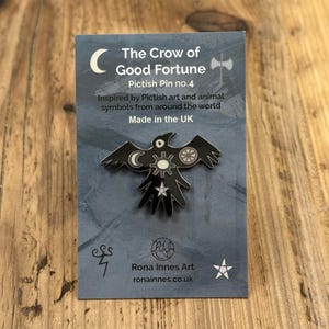 May include: A black enamel pin depicting a crow with a crescent moon, gear, and wheel symbols. The pin is displayed on a grey card with the text "The Crow of Good Fortune" and "Pictish Pin no. 4". The card also states "Made in the UK".
