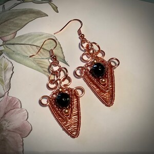 May include: A pair of copper wire earrings with black gemstone accents. The earrings are shaped like a teardrop with a wire design and a black gemstone in the centre.