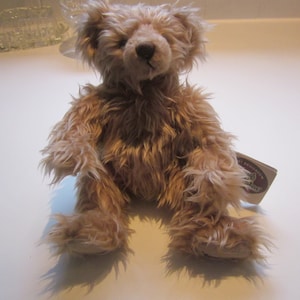 May include: A light brown teddy bear with a fluffy coat, sitting on a white surface. The bear has a small tag attached to its right leg.