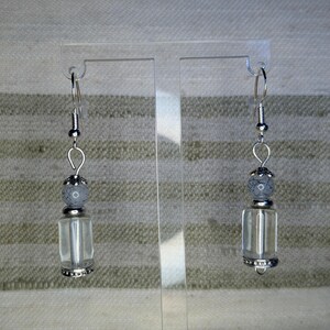 May include: A pair of dangle earrings with clear, rectangular glass vials. Each earring features a gray, speckled bead above the vial and a decorative silver cap and base. The earrings hang from silver-colored fish hook ear wires.