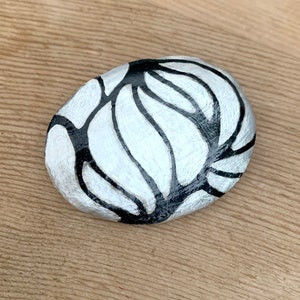 以下が含まれることがあります： 黒と銀の模様で描かれた、滑らかで楕円形の石。模様は、流れるような有機的なパターンを生み出す、一連の曲線で構成されています。