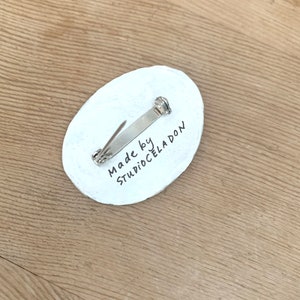 以下が含まれることがあります： 銀色のピン留めが付いた白い楕円形のピン。ピンには「Made by StudioCeladon」という黒い文字が書かれています。
