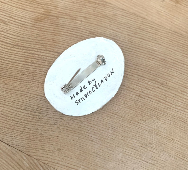 以下が含まれることがあります： 銀色のピン留めが付いた白い楕円形のピン。ピンには「Made by StudioCeladon」という黒い文字が書かれています。
